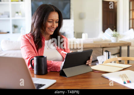 Smiling middle aged woman sitting at the table in her dining room drawing on a tablet computer with a stylus, close up Stock Photo