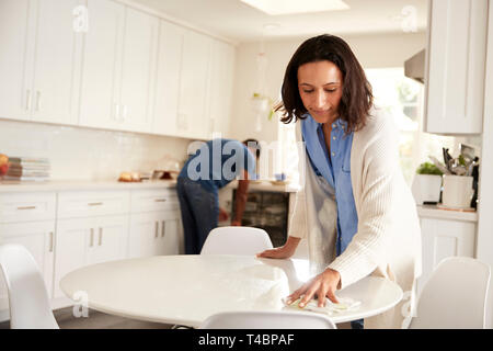 Millennial woman cleaning kitchen table while her partner also does chores in the background, focus on foreground Stock Photo