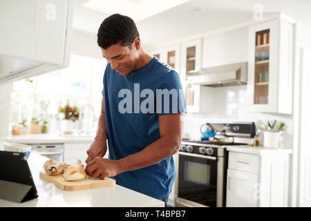 Side view of adult man standing at kitchen worktop preparing food using recipe on a tablet computer, close up, selective focus Stock Photo