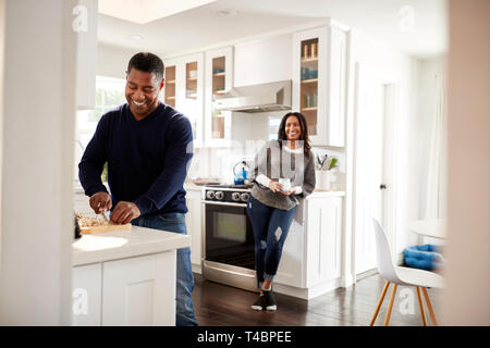 Middle aged man standing at worktop in the kitchen preparing food, his partner leaning on the kitchen cabinet behind him talking, selective focus