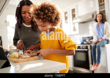 Grandmother helping her granddaughter prepare food in the kitchen, mother sitting in the background, focus on foreground Stock Photo