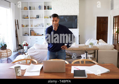 Millennial Hispanic man standing at table in the dining room looking down at his laptop computer screen Stock Photo