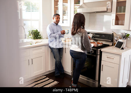 Middle aged mixed race couple standing together in the kitchen preparing food on the hob using a recipe on a tablet computer, seen from doorway Stock Photo