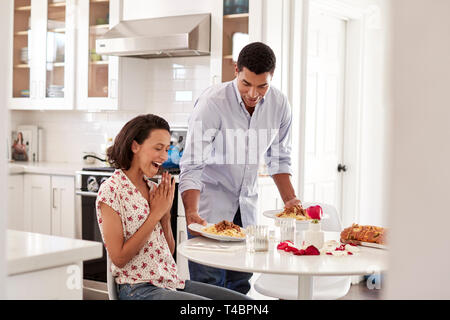Young mixed race adult woman sitting at the table in the kitchen, her partner surprising her by serving a romantic meal, selective focus