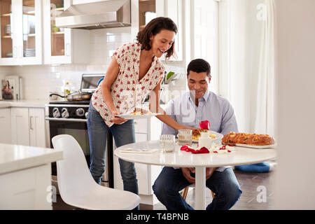 Young adult man sitting at the table in the kitchen is served a romantic meal by his partner, selective focus Stock Photo