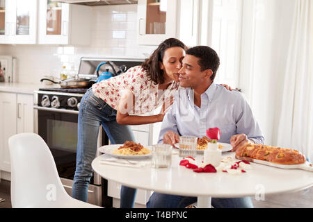 Young adult woman kissing her partner, sitting at the table in their kitchen for a romantic dinner, selective focus Stock Photo