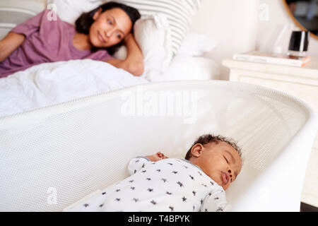Close up of young adult mother lying on her bed looking down at her three month old baby sleeping in his cot