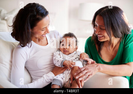 Millennial mixed race mother sitting in an armchair holding her three month old baby son, her mother kneeling beside them Stock Photo
