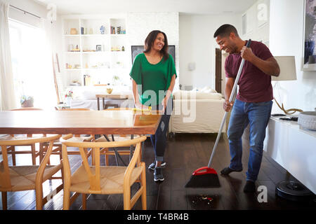 Millennial man sweeping the floor in the dining room while his partner stands talking to him Stock Photo