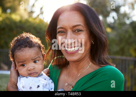 Happy mixed race middle aged grandmother holding her three month old grandson in the garden and smiling to camera, close up, head and shoulders Stock Photo