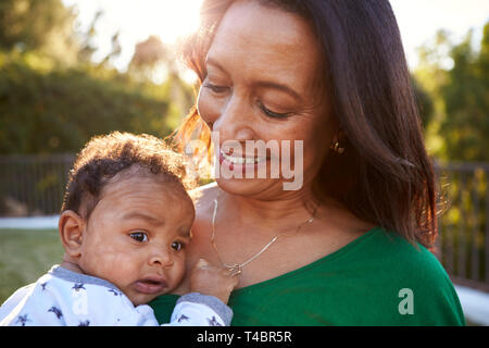 Happy mixed race middle aged grandmother holding her three month old grandson in the garden, close up, head and shoulders Stock Photo
