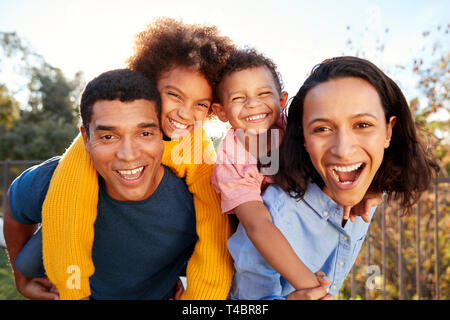 Young mixed race parents parents having fun piggybacking their children in the garden, looking to camera and laughing, close up Stock Photo