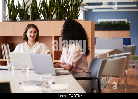 Two Businesswomen Working On Laptops Sitting At Table In Modern Open Plan Office Stock Photo