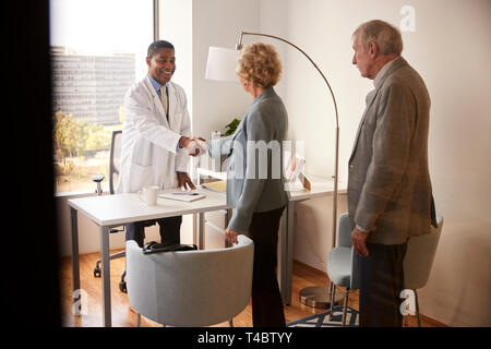 Senior Couple Being Greeted By Male Doctor With Handshake On Visit To Hospital For Consultation Stock Photo