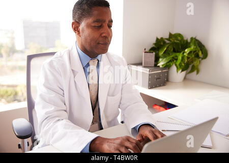 Male Doctor Wearing White Coat In Office Sitting At Desk Working On Laptop Stock Photo