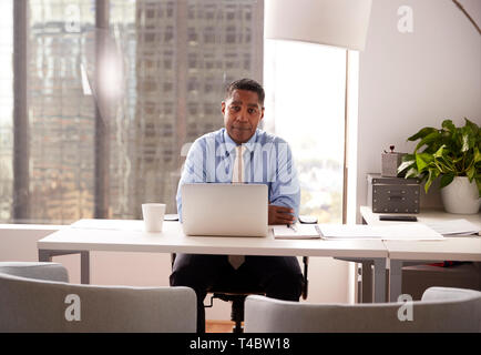 Portrait Of Male Financial Advisor In Modern Office Sitting At Desk Working On Laptop Stock Photo