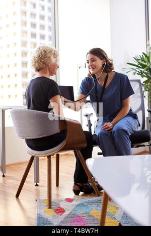 Female Doctor Wearing Scrubs In Office Listening To Senior Female Patients Chest Using Stethoscope Stock Photo
