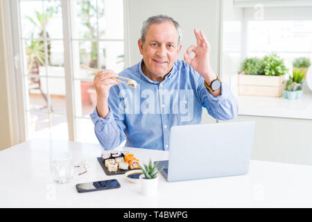 Handsome business senior man eating delivery sushi while working using laptop doing ok sign with fingers, excellent symbol Stock Photo