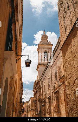 Beautiful view of narrow medieval street in Mdina with Carmelite Church Bell Tower an blue sky with clouds at the background. Ancient capital of Malta Stock Photo