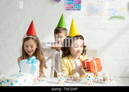 adorable kids in party caps sitting at table with gift boxes during birthday party at home Stock Photo