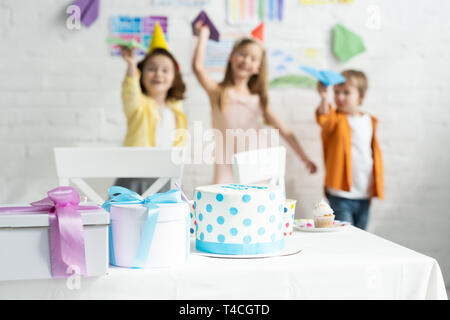 selective focus of cake and presents on table and kids playing with paper planes during birthday party Stock Photo