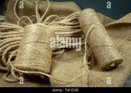 Two spools of burlap threads or jute twine and coil of linen rope on sackcloth fabric in close-up on grey background Stock Photo