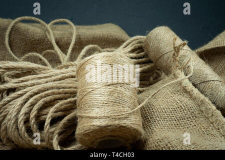 Two spools of burlap threads or jute twine and coil of linen rope on sackcloth fabric in close-up on grey background Stock Photo