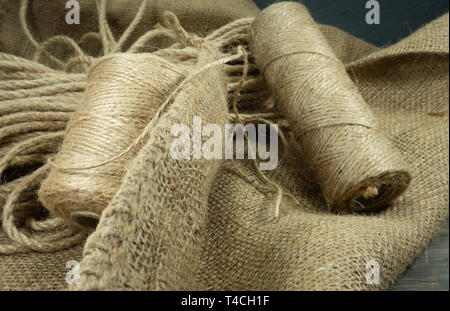 Two spools of burlap threads or jute twine and coil of linen rope on sackcloth fabric in close-up on grey background Stock Photo