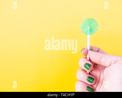 A woman with green painted nails with a green lollipop in her hand and yellow background Stock Photo