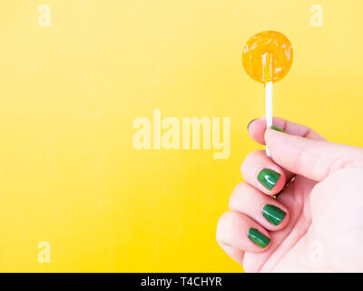 A woman with green painted nails with an orange lollipop in her hand and yellow background Stock Photo