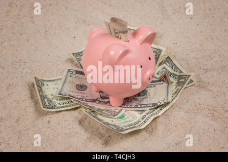 Horizontal shot of a pink piggy bank sitting on a pile of money with money in its slot all on a sand beach background. Stock Photo