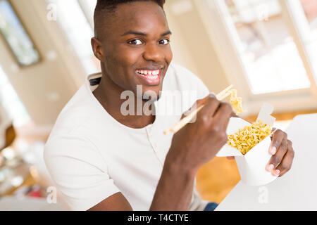 Handsome african man eating asian noodles in a delivery box, smiling enjoying lunch using chopsticks Stock Photo