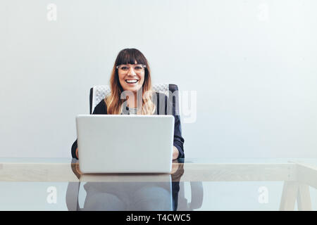 Smiling businesswoman sitting at her desk in office. Cheerful woman working on laptop computer sitting at her desk. Stock Photo