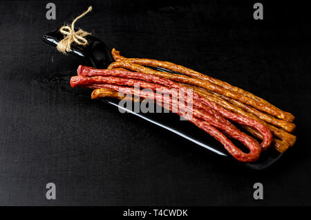 Long thin smoked sausages on black background on a plate made from bottle. Traditional Polish Kabanosy or Frisian dried sausage sticks. Snaks for beer Stock Photo