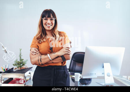 Portrait of a woman entrepreneur standing in office with arms crossed. Smiling businesswoman in eyeglasses standing near her desk in office. Stock Photo