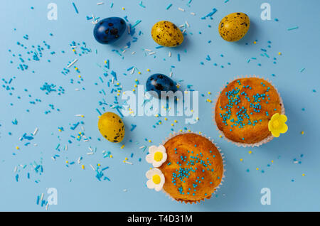 Muffins decorated with sprinkles, flowers of mastic and yellow and blue easter quail eggs on pastel blue background. Top view. Easter Holidays and gre Stock Photo