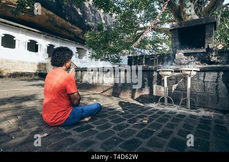 Young man (Buddhist) praying against ancient cave temple complex. Dambulla, Sri Lanka. Stock Photo