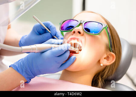 Dental hygienist performing teeth cleaning procedure to elementary age girl in pediatric dental clinic. Doctor using prophy brush on handpiece to remo Stock Photo