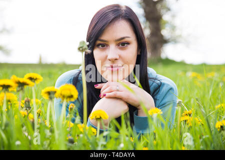 Beautiful happy young woman lying on the field in green grass and smelling yellow dandelion, enjoying nature. Freedom and nature concept. Stock Photo