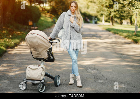 stylish young blonde mother pushing around a stroller with baby inside in a sunny park Stock Photo