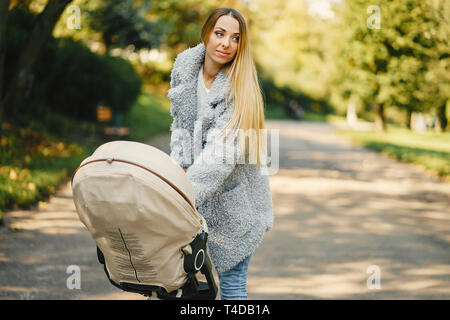 stylish young blonde mother pushing around a stroller with baby inside in a sunny park Stock Photo