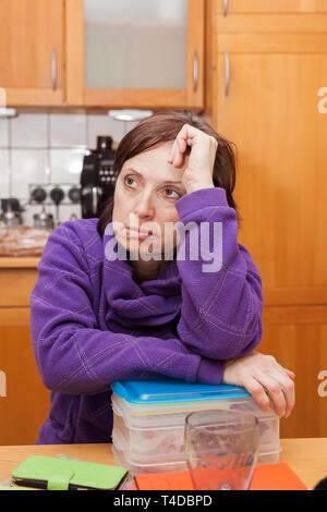 Middle-aging woman is sitting undecided in the kitchen. Stock Photo