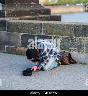 Cold Homeless man on his knees begging for money from tourists on the Charles Bridge in Prague - Spring 2019 Stock Photo