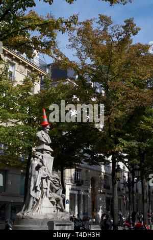 A statue on Place de la Sorbonne, Paris, France. Stone statue of a man, under trees in front of buildings. A red and silver / grey striped traffic con
