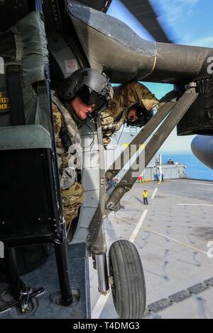 Two U.S. Army UH-60 Black Hawk helicopter crew members, assigned to the 1-108th Assault Helicopter Battalion, Kansas Army National Guard, survey the landing zone in preparation to land aboard a U.S. Navy ship in the Arabian Gulf, March 29, 2019.  The 1-108th AHB deployed to the Middle East to complete aviation missions in support of Operations Spartan Shield and Inherent Resolve. Stock Photo