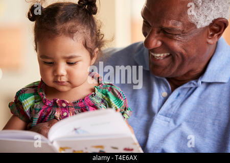 Grandfather Sitting On Sofa At Home With Baby Granddaughter Reading Book Together Stock Photo