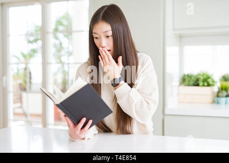 Beautiful Asian woman reading a book cover mouth with hand shocked with shame for mistake, expression of fear, scared in silence, secret concept Stock Photo