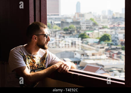 Bearded young man with sunglasses looking out of Golden Mount Wat Saket temple window over Bangkok, Thailand. Stock Photo