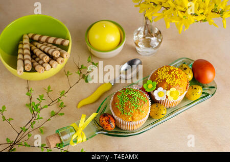 Muffins decorated with sprinkles, flowers of mastic, colored easter quail eggs on a kitchen table. Top view. Happy Easter holiday and greeting card co Stock Photo