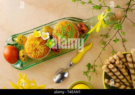 Muffins decorated with sprinkles, flowers of mastic, colored easter quail eggs on a kitchen table. Top view. Happy Easter holiday and greeting card co Stock Photo